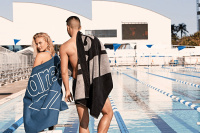 Produkten är handduken till höger i en annan färg En dam och en man med varsin Arena handduk framför en utomhuspool