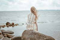 En dam i en morgonrock sitter på en sten vid havet med ryggen mot kameran