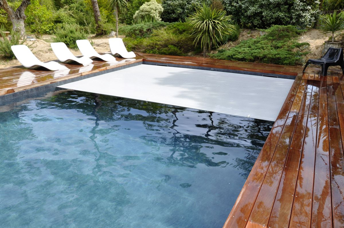 Rollinside Lamelltäcke delvis inrullat i en pool med en trädgård i bakgrunden