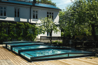 Pooltak Leia Compact 3 x 6 m Pooltaket över en utomhuspool med träsarg och ett hus i bakgrunden