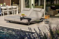 Dubbelmodell med extra tillbehör En dubbel solsäng i grått med nackkuddar står vid en pool. En liten skål med frukt och en handduk ligger på bordet mellan sängarna.