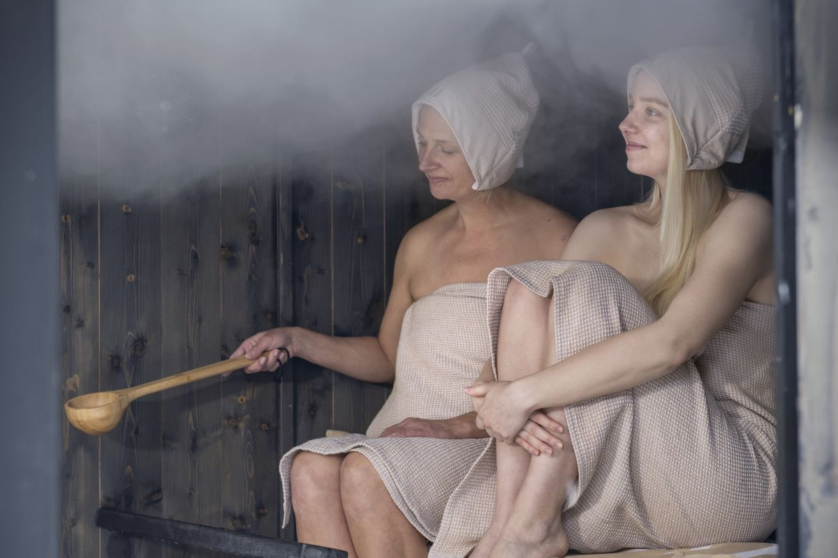 Två kvinnor sitter i en bastu och en håller i skopan