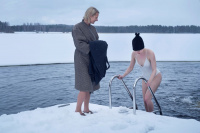 En dam klättrar upp ur en isvak klädd i bastumössan medan en annan väntar på bryggan