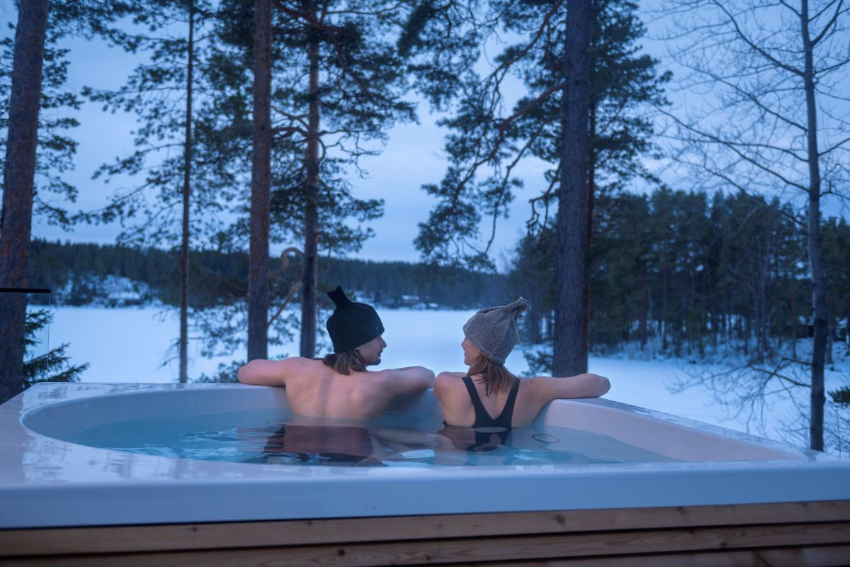 Ett par med varsin bastumössa sitter i ett spabad utomhus med ryggen mot kameran